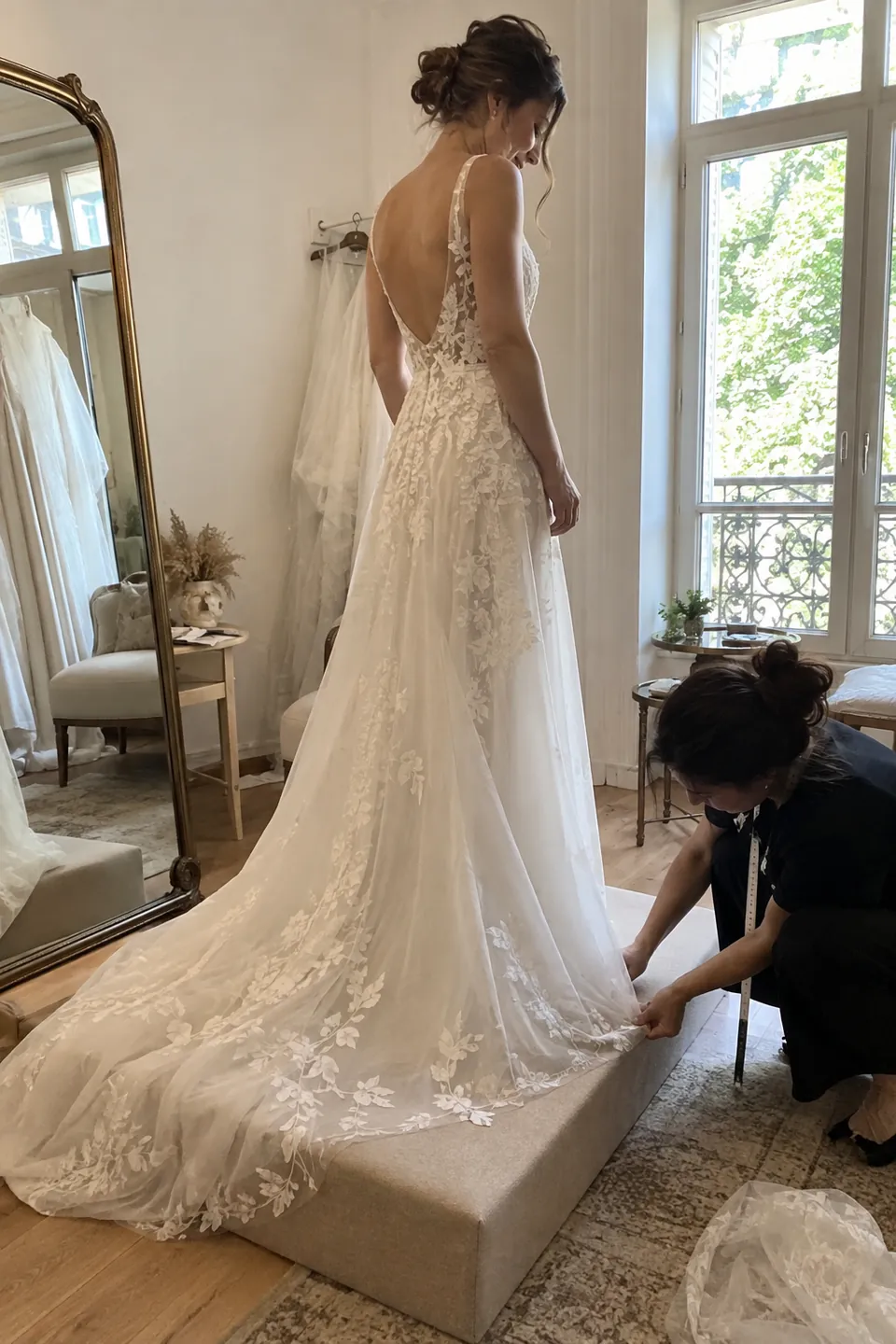 A bride standing on a low platform while a tailor marks the hem of a long lace gown