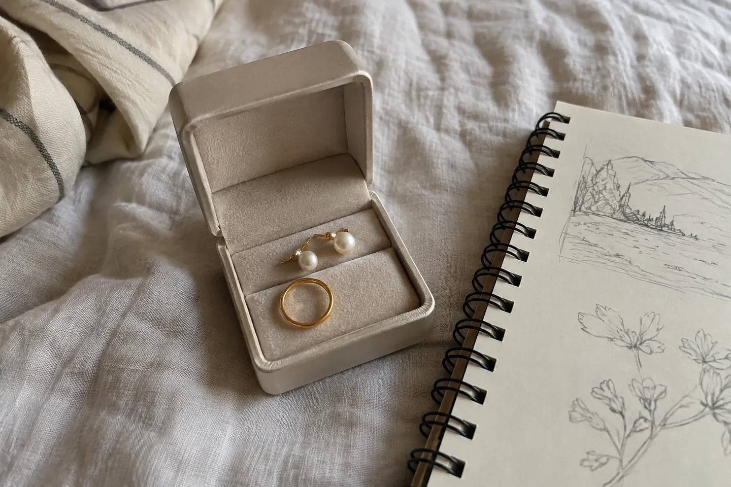 A small velvet box with pearl earrings and a plain gold ring beside a sketchbook